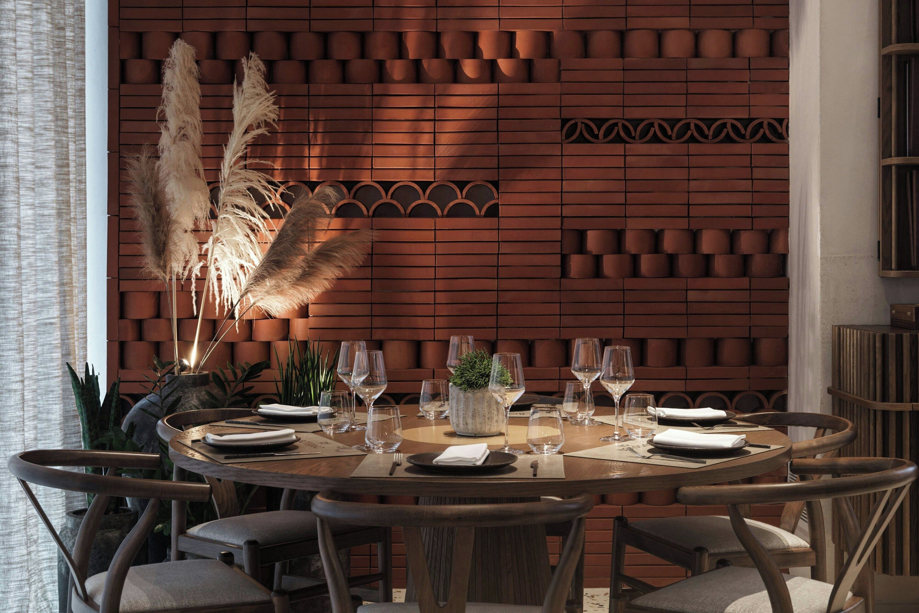 A round dining table set with plates, glasses, and cutlery, surrounded by chairs, with a brick wall and decorative plants in the background.