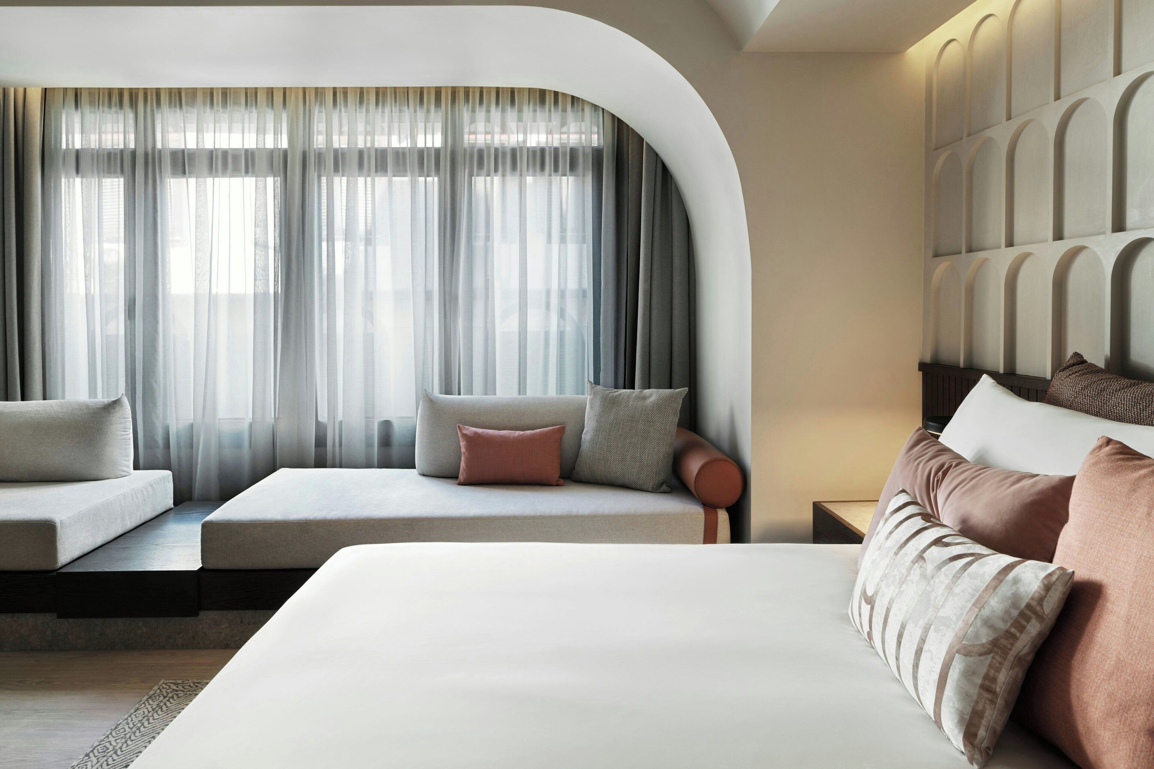 Una habitación de hotel moderna con una cama grande, un sofá y un ventanal curvo con cortinas translúcidas.