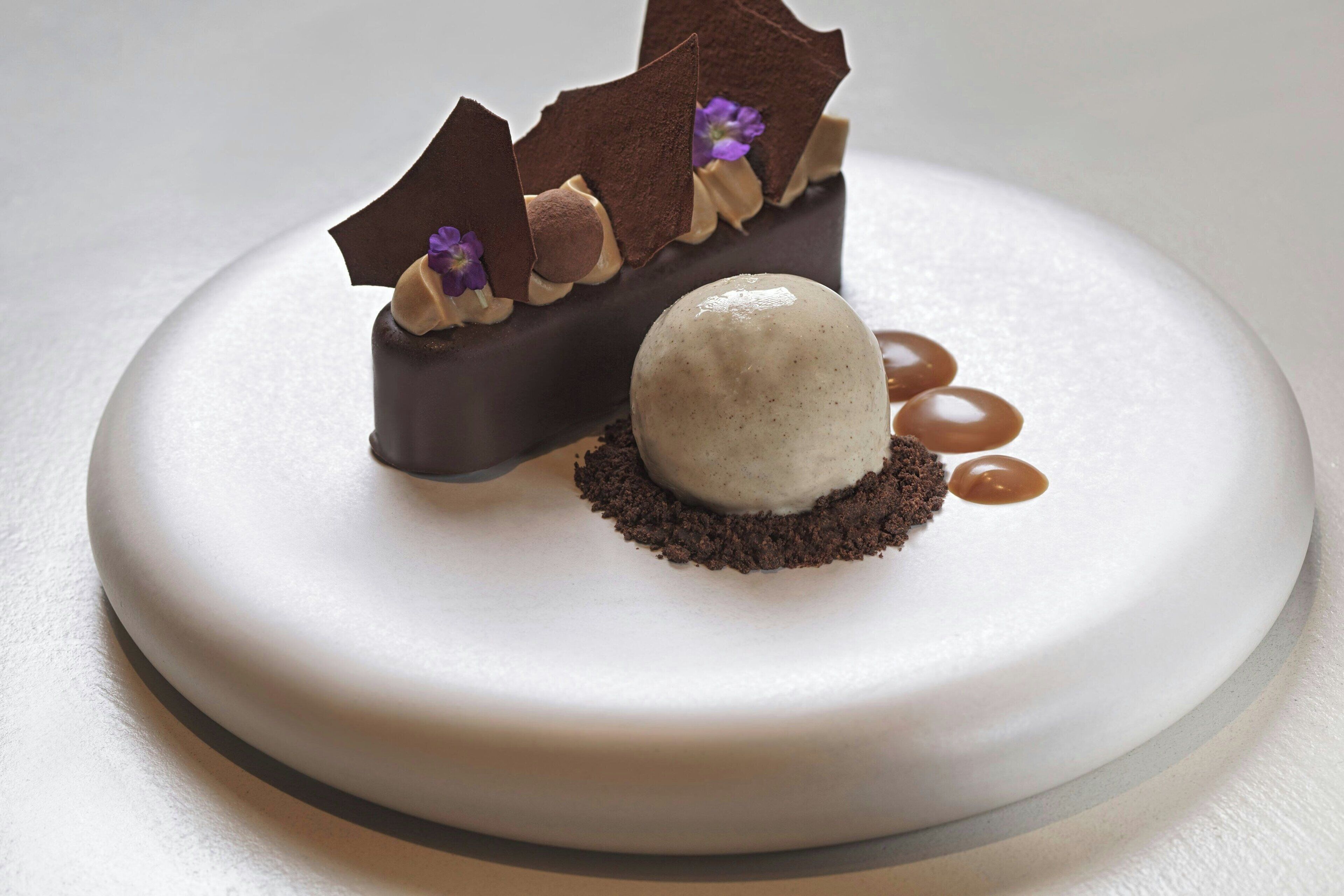 Un postre con un trozo de chocolate, una bola de helado y un hilo de caramelo sobre un plato blanco.