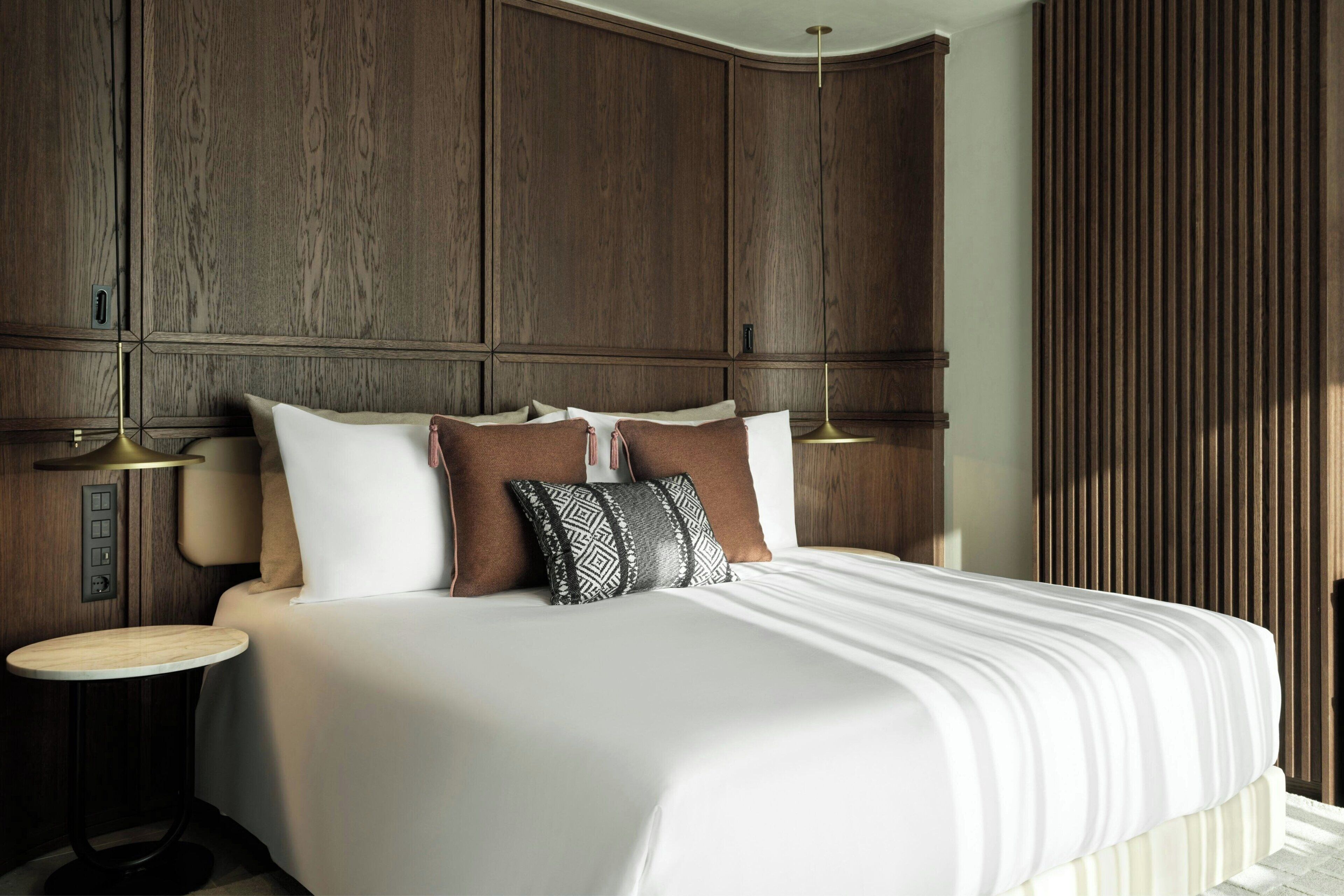 Una cama bien hecha con sábanas blancas y varias almohadas, una mesita de noche con lámpara y un cabecero de madera.