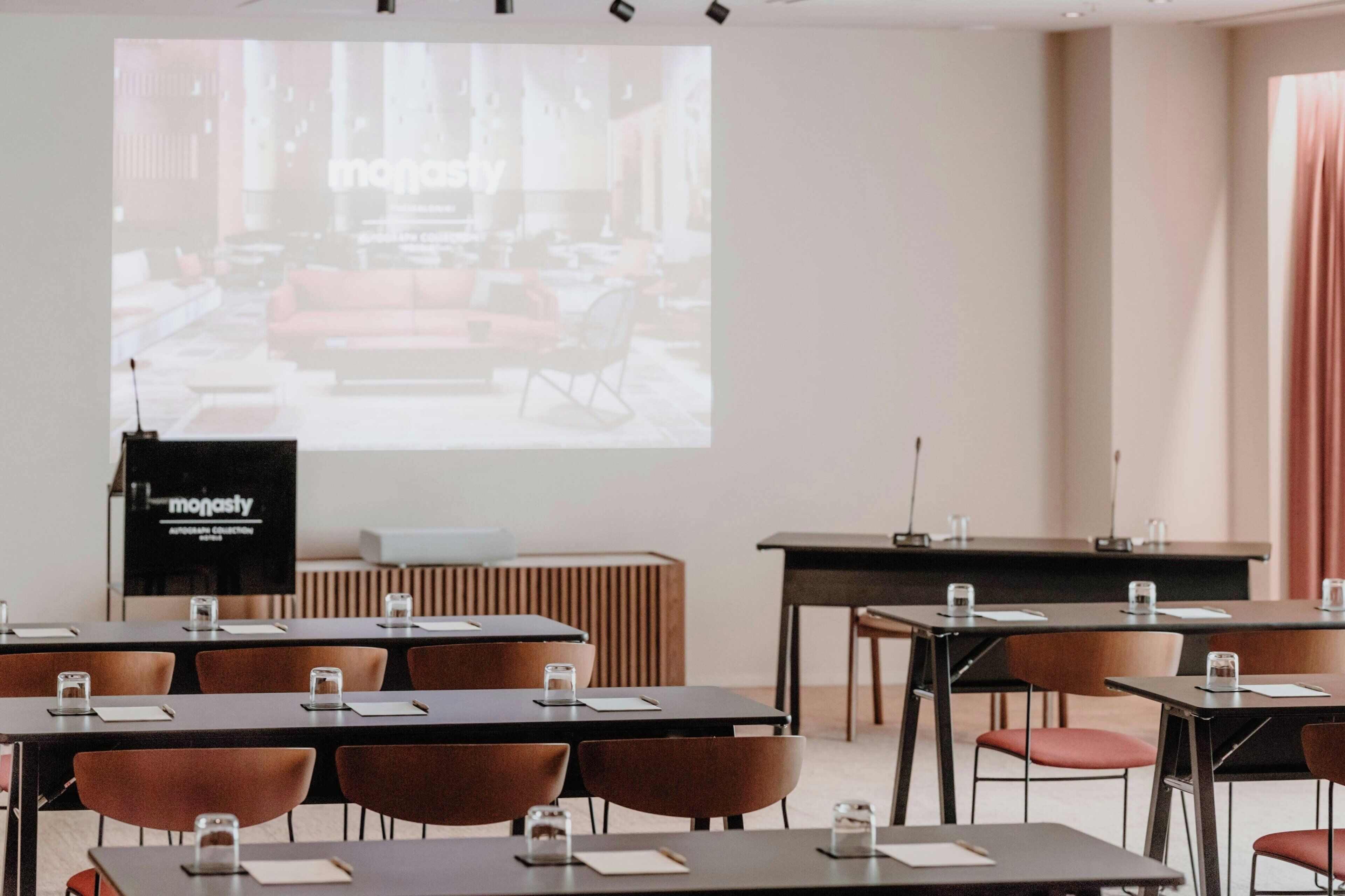 Un salón de conferencias con mesas y sillas, una pantalla de proyector mostrando una habitación, y un atril con la palabra "monasty".