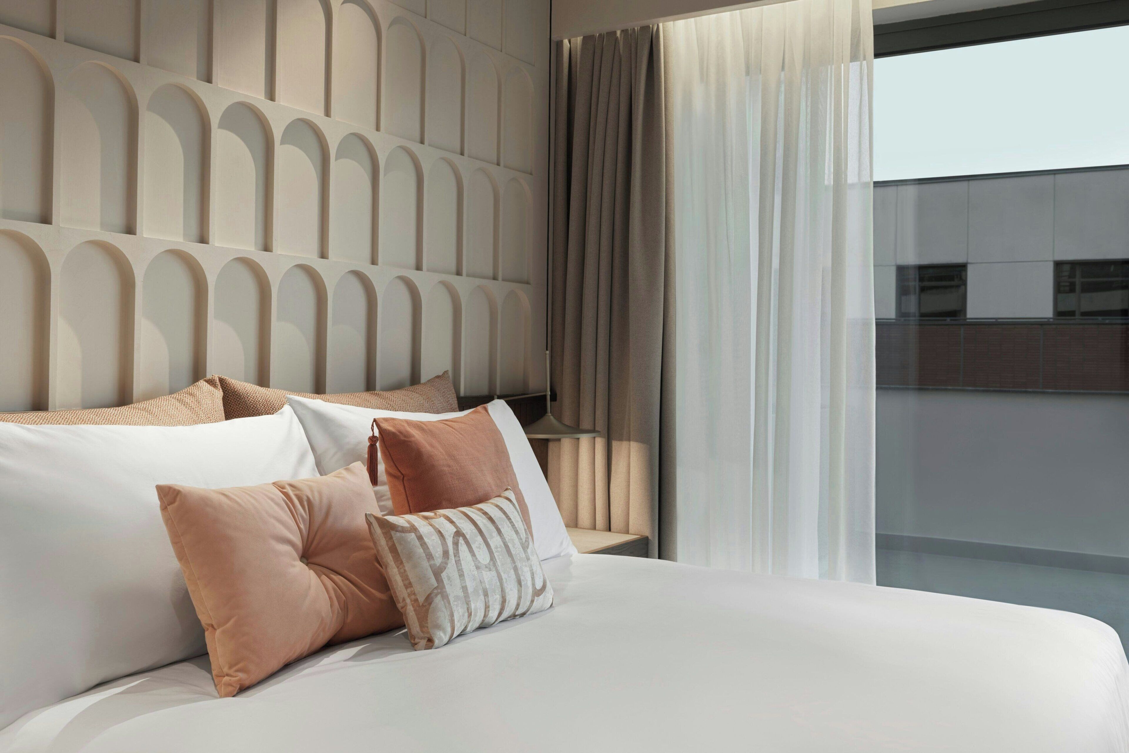 A modern hotel room with a large bed, a headboard with a geometric pattern, and a view of the city through large windows.