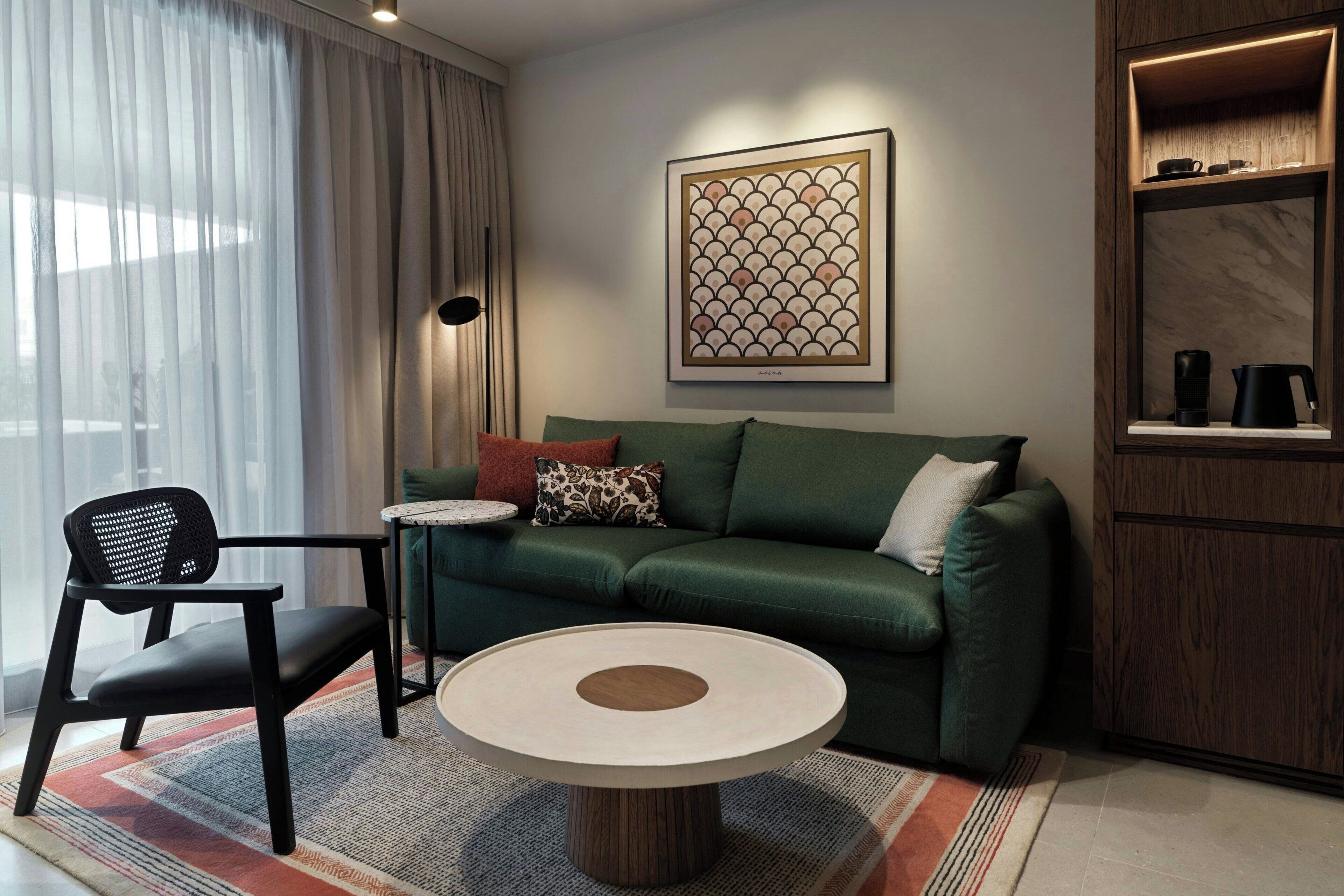 Uma sala de estar moderna com um sofá verde, uma mesa de centro redonda, uma cadeira preta, uma pequena mesa lateral e uma obra de arte emoldurada na parede.