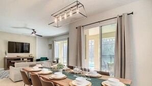 Een eetruimte met een lange tafel gedekt voor zes personen, een televisie op de achtergrond en uitzicht op het buitenleven door grote ramen.