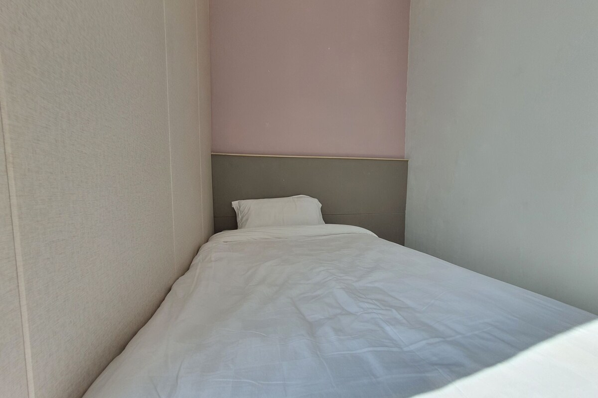 Een kleine kamer met een enkel bed, een eenvoudig hoofdeinde en kaal stucwerk.