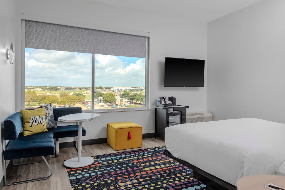 A hotel room with a bed, a sofa, a small table, a yellow ottoman, a TV, and a window with a city view.