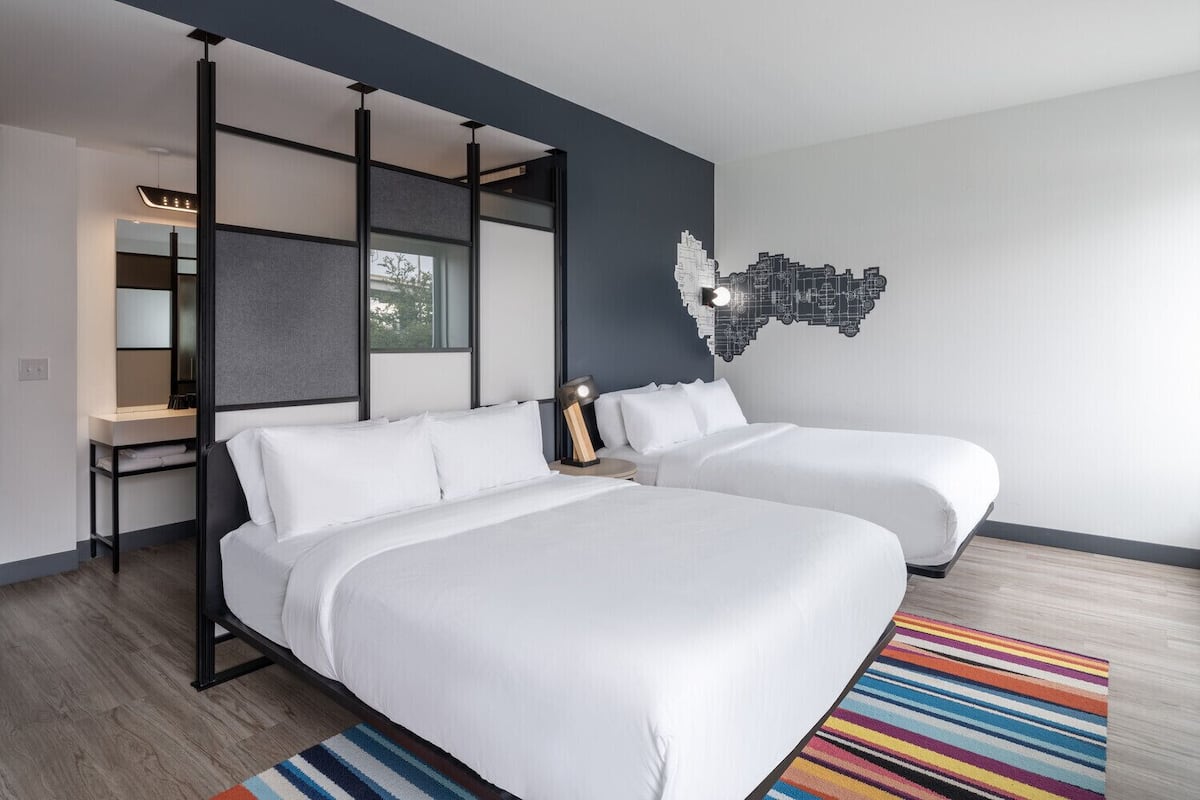 A modern hotel room with two beds, a desk, and a cityscape wall art.