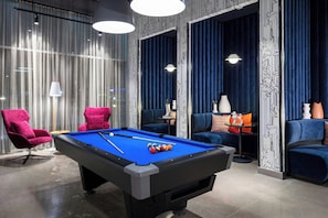 Ein Billardtisch mit Kugeln, ein roter Sessel und ein blaues Sofa in einem Raum mit gemustertem Wandpapier und blauen Vorhängen.