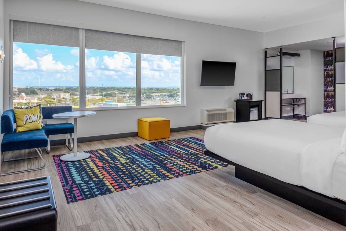 A modern hotel room with a large bed, a TV mounted on the wall, a small table, a blue chair, a colorful rug, and a view of the city through large windows.
