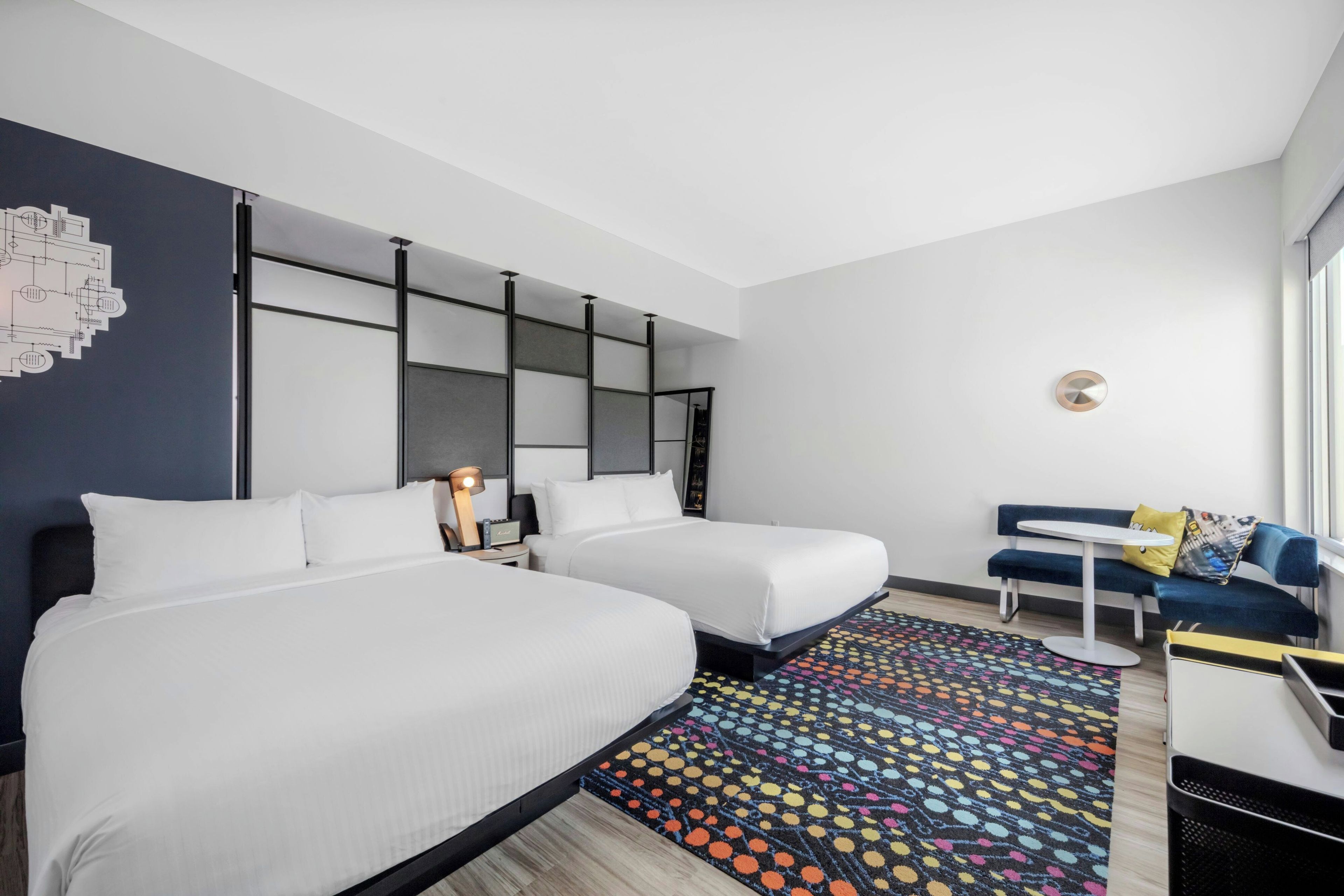 Quarto de hotel com duas camas, uma escrivaninha, uma cadeira, um tapete colorido e parede com design geométrico.