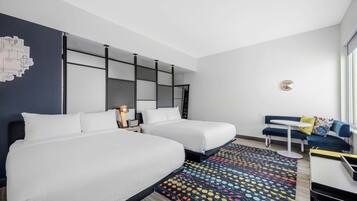 Quarto de hotel com duas camas, uma escrivaninha, uma cadeira, um tapete colorido e parede com design geométrico.