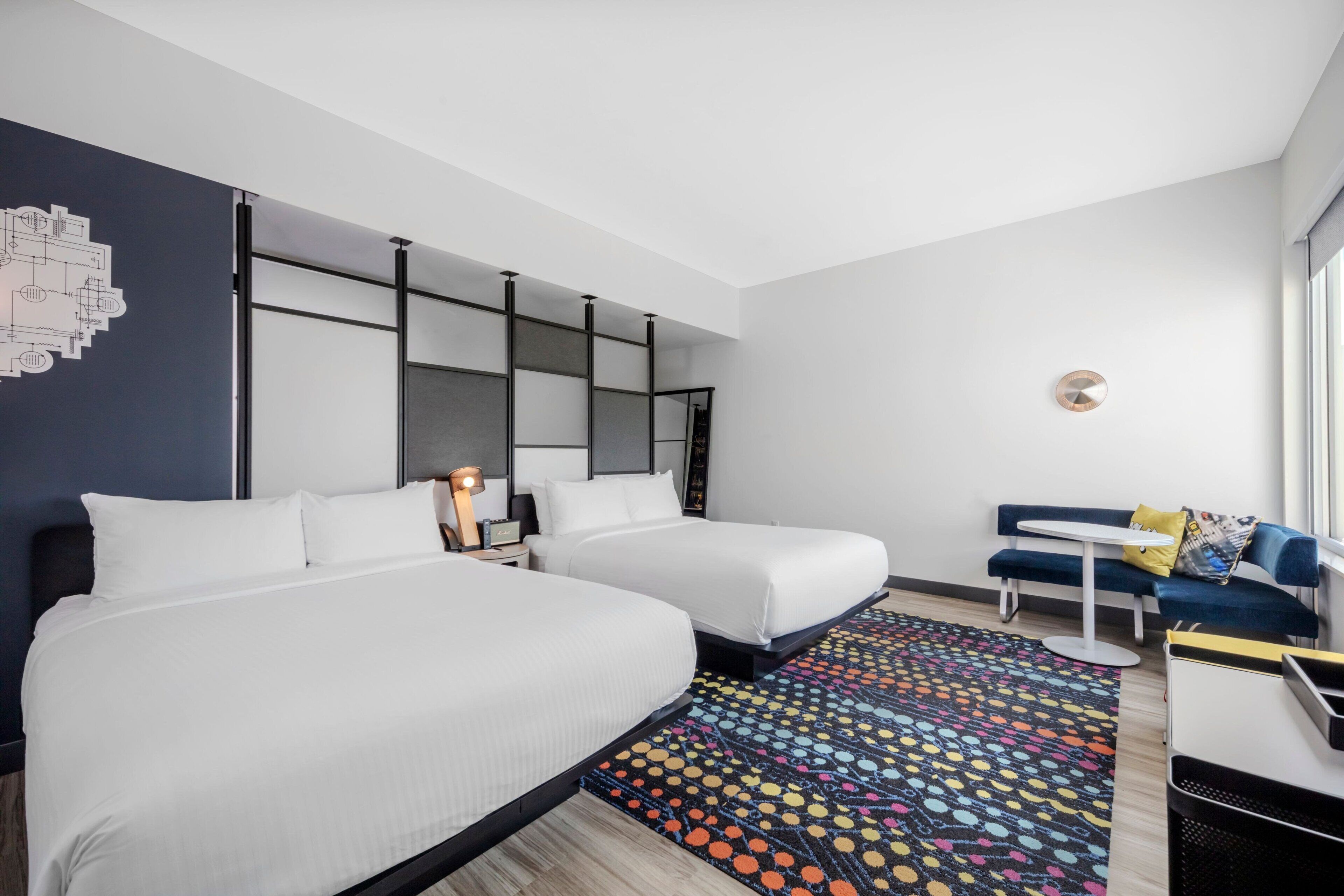 Habitación de hotel con dos camas, un escritorio, una silla, un tapete colorido y una lámpara fijada a la pared.