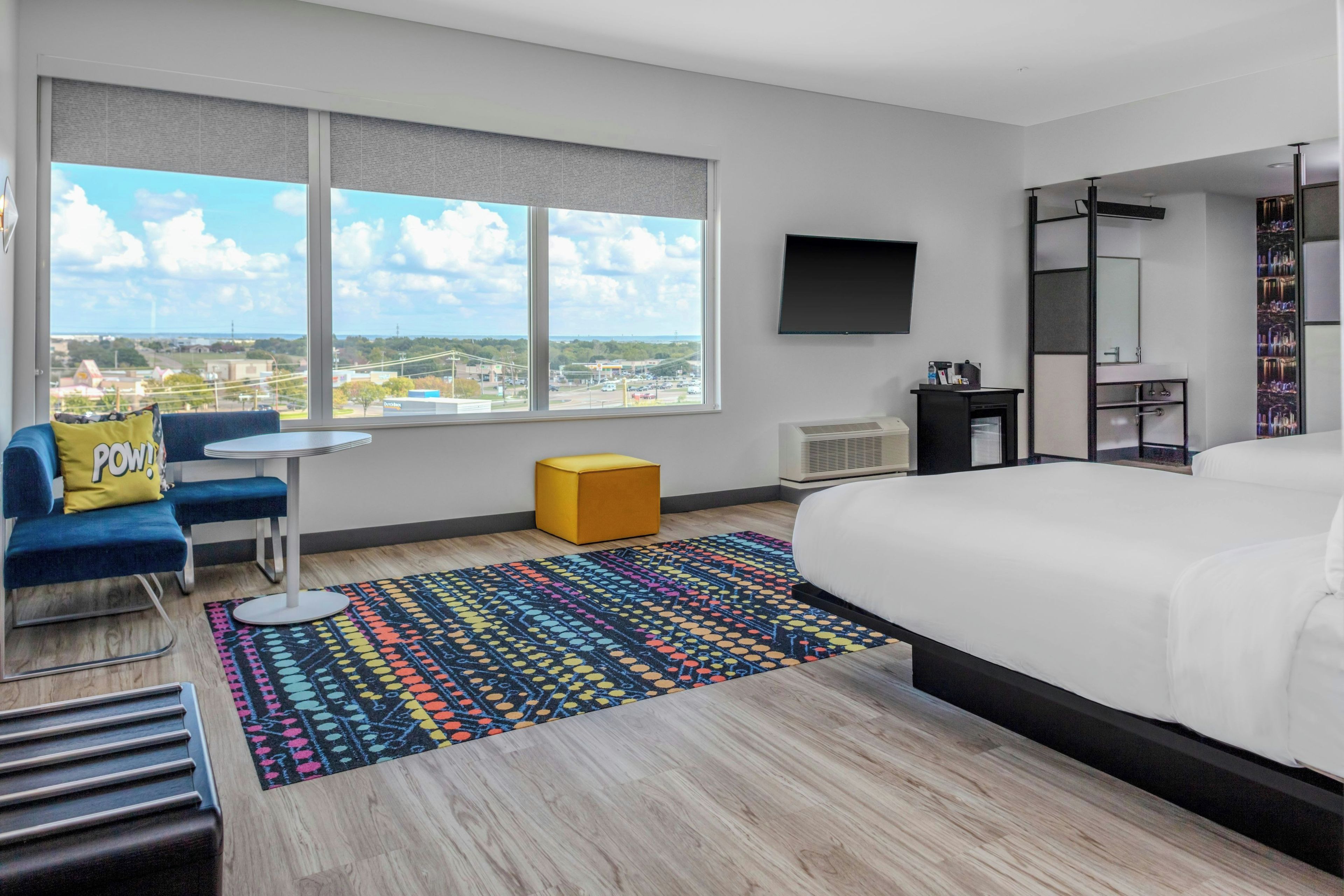 A modern hotel room with a large bed, a desk, a chair, a colorful rug, and a view of the city.