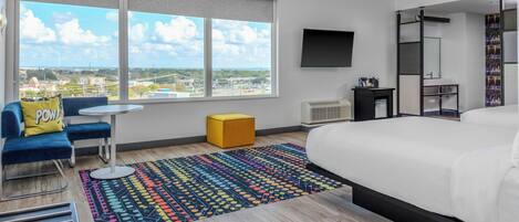 A modern hotel room with a large bed, a desk, a chair, a colorful rug, and a view of the city.