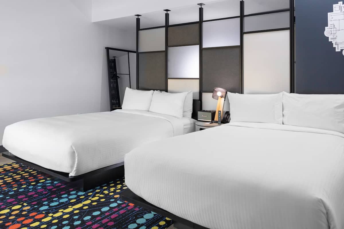 A hotel room with two beds, a colorful rug, a nightstand with a lamp, and a wall with a geometric design.