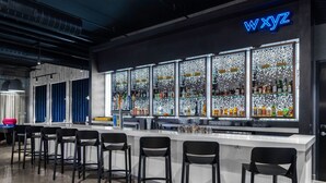 Un espace bar agrémenté d’un panneau néon indiquant « WXYZ », de plusieurs étagères remplies de bouteilles et de tabourets de bar noirs.