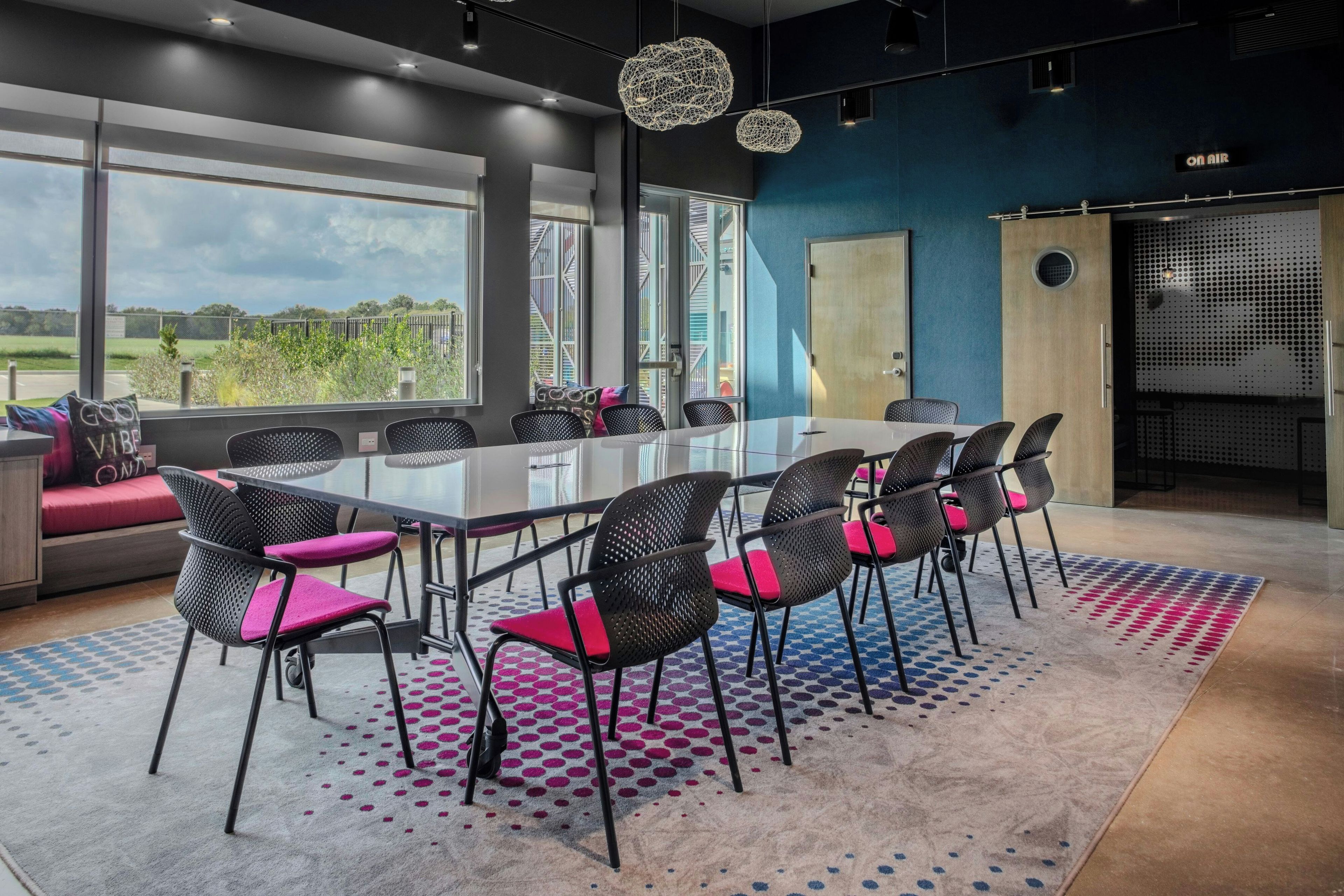 Una sala de conferencias moderna con una mesa de vidrio, sillas negras con cojines rosados y vistas al exterior.