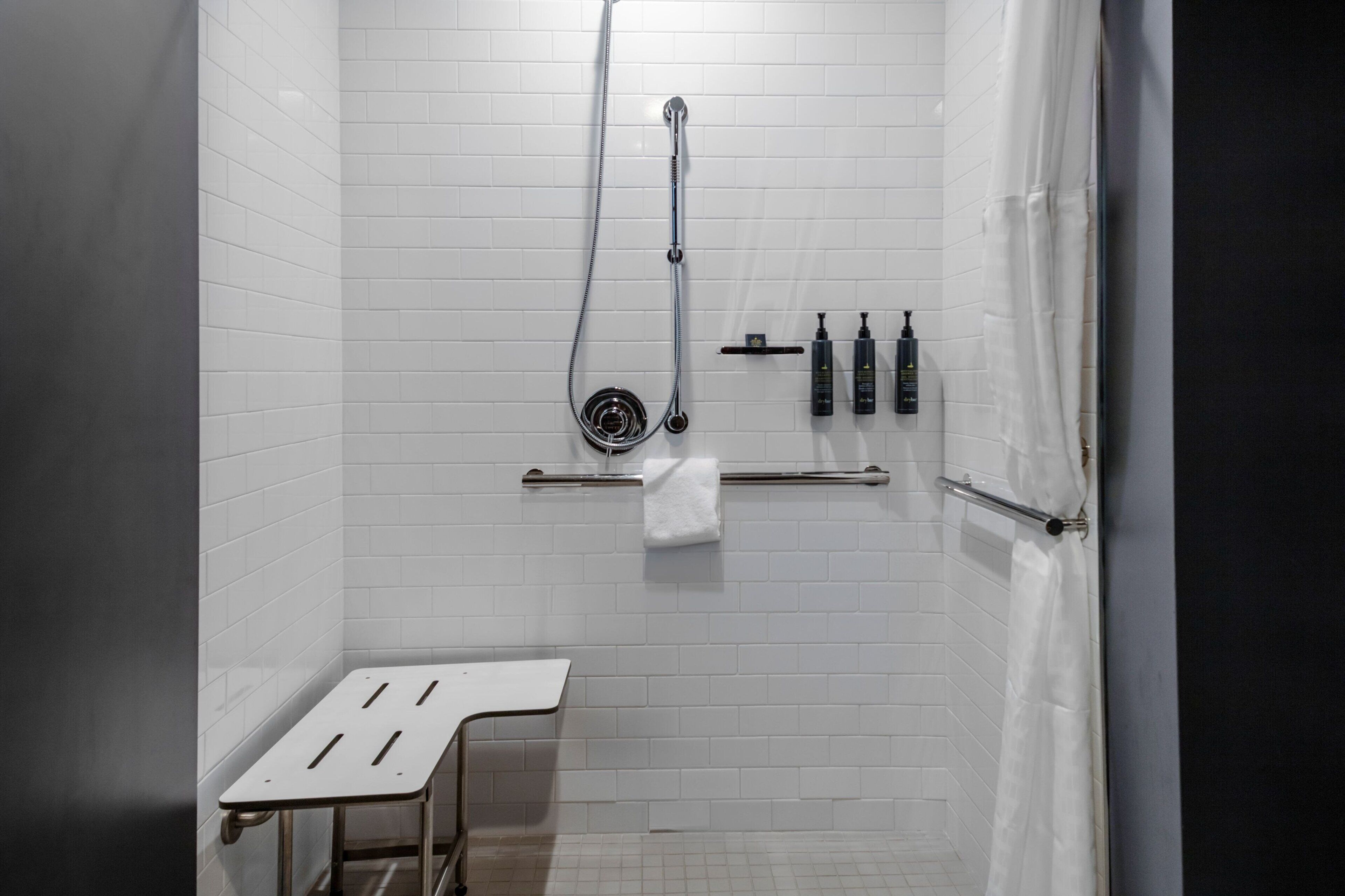 A modern bathroom with white tiled walls, a shower with a handheld showerhead, a towel rack, and a small table.