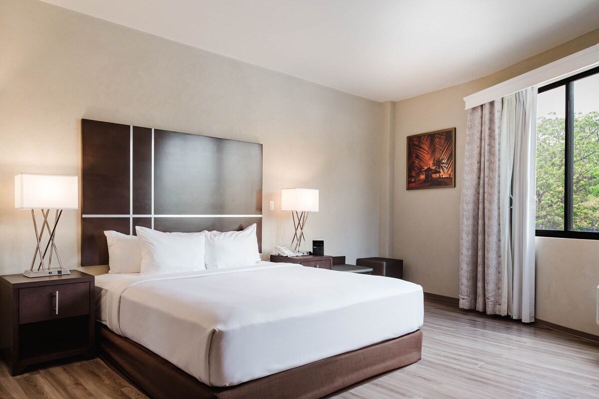 A hotel room with a large bed, two bedside tables with lamps, a nightstand, a painting, and a window with curtains.