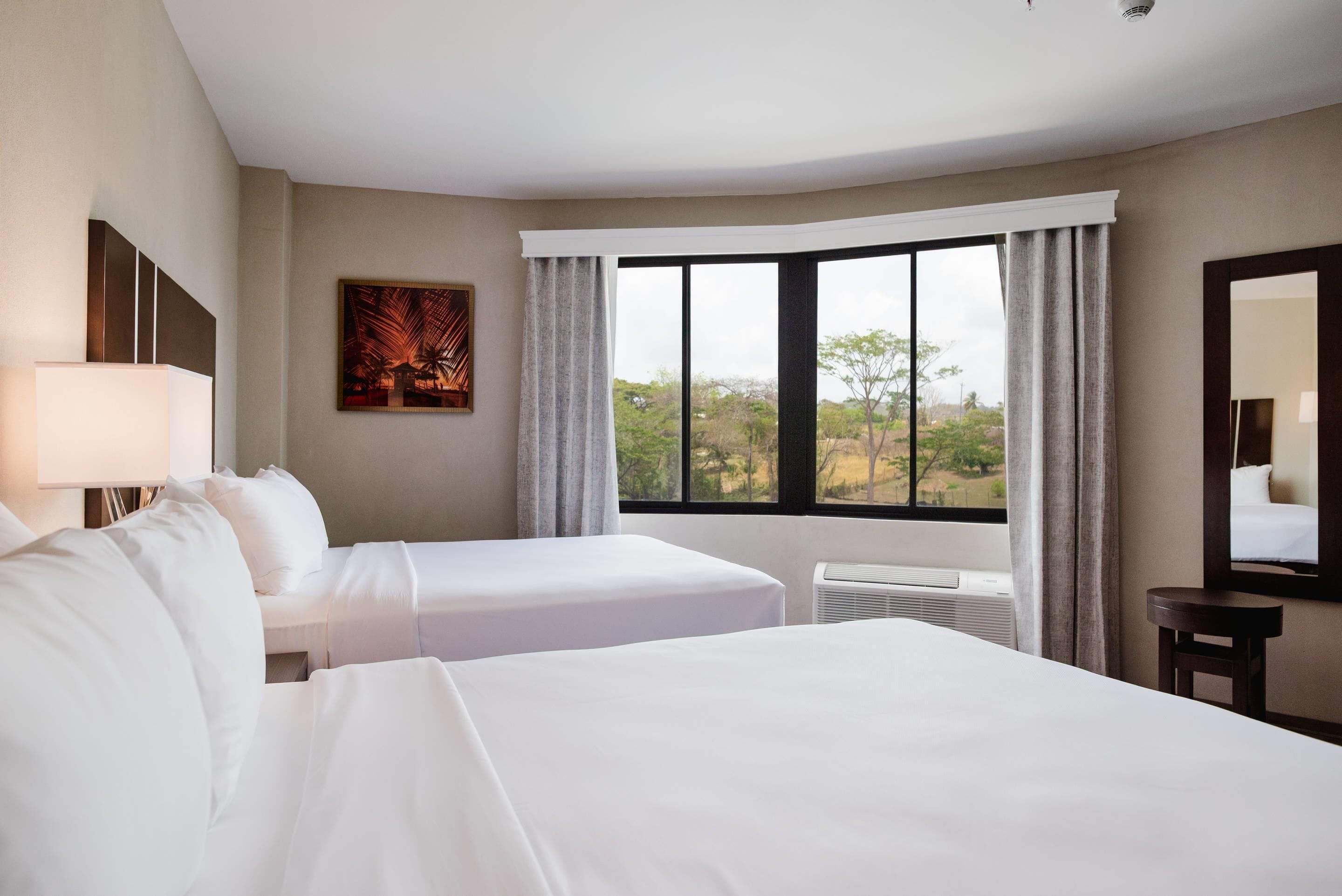 A hotel room with two beds, a large window with curtains, a painting on the wall, and a mirror.