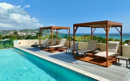 A poolside area with lounge chairs and wooden gazebos, overlooking a scenic view of the ocean and a distant town.
