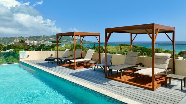 A poolside area with lounge chairs and wooden gazebos, overlooking a scenic view of the ocean and a distant town.