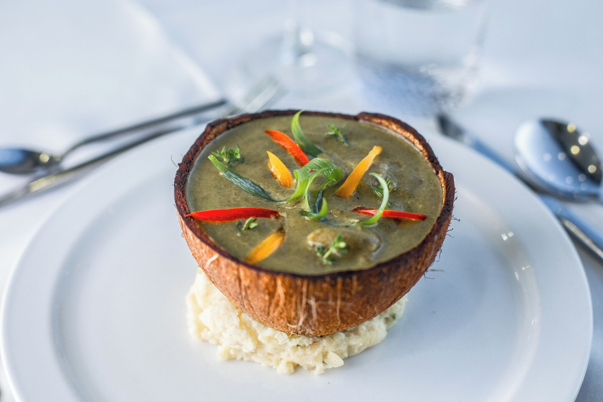 A dish with a coconut shell bowl containing a soup with vegetables, served on a white plate.