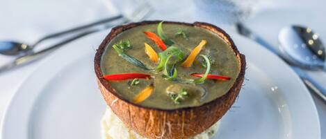 A dish with a coconut shell bowl containing a soup with vegetables, served on a white plate.