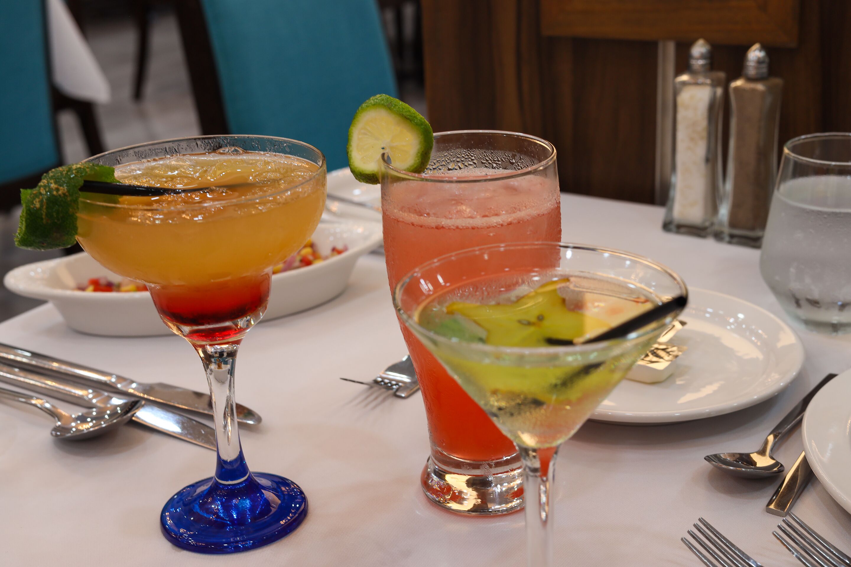 A table with two cocktails, a fork, a spoon, and a glass of water.