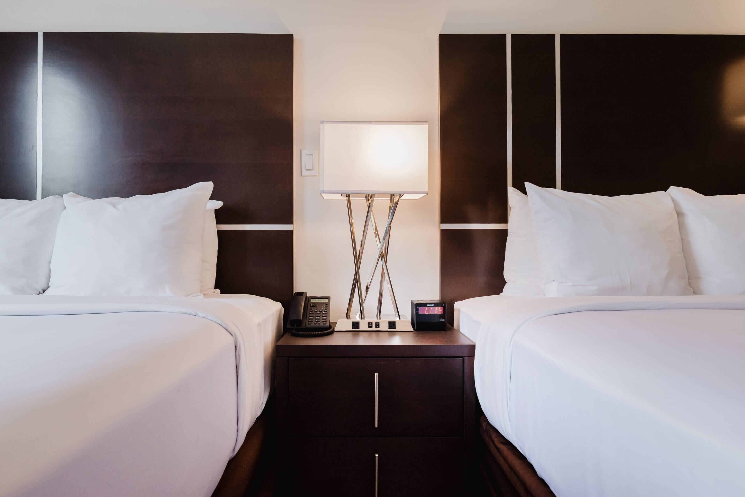Una camera d'albergo con due letti, un comodino con lampada, un telefono e una sveglia digitale.