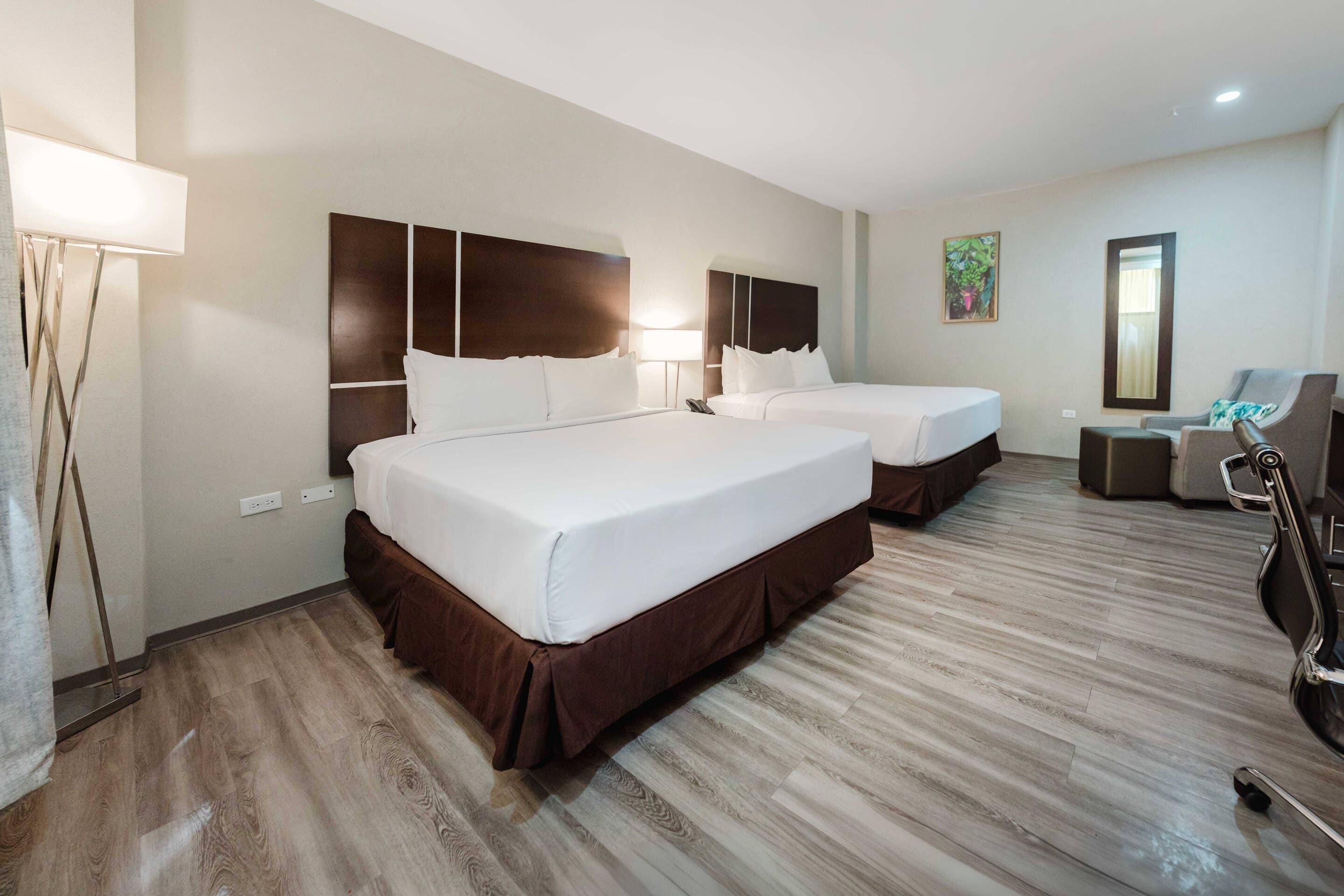 A hotel room with two beds, a desk, a chair, and a lamp.
