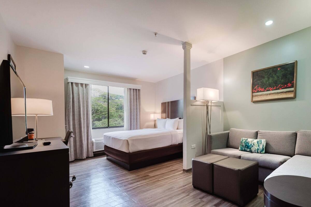 A hotel room with a bed, a sofa, a desk with a lamp, and a painting on the wall.