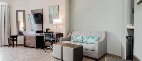 A hotel room with a flat-screen TV, a desk with a chair, a sofa with a patterned cushion, and a small table.