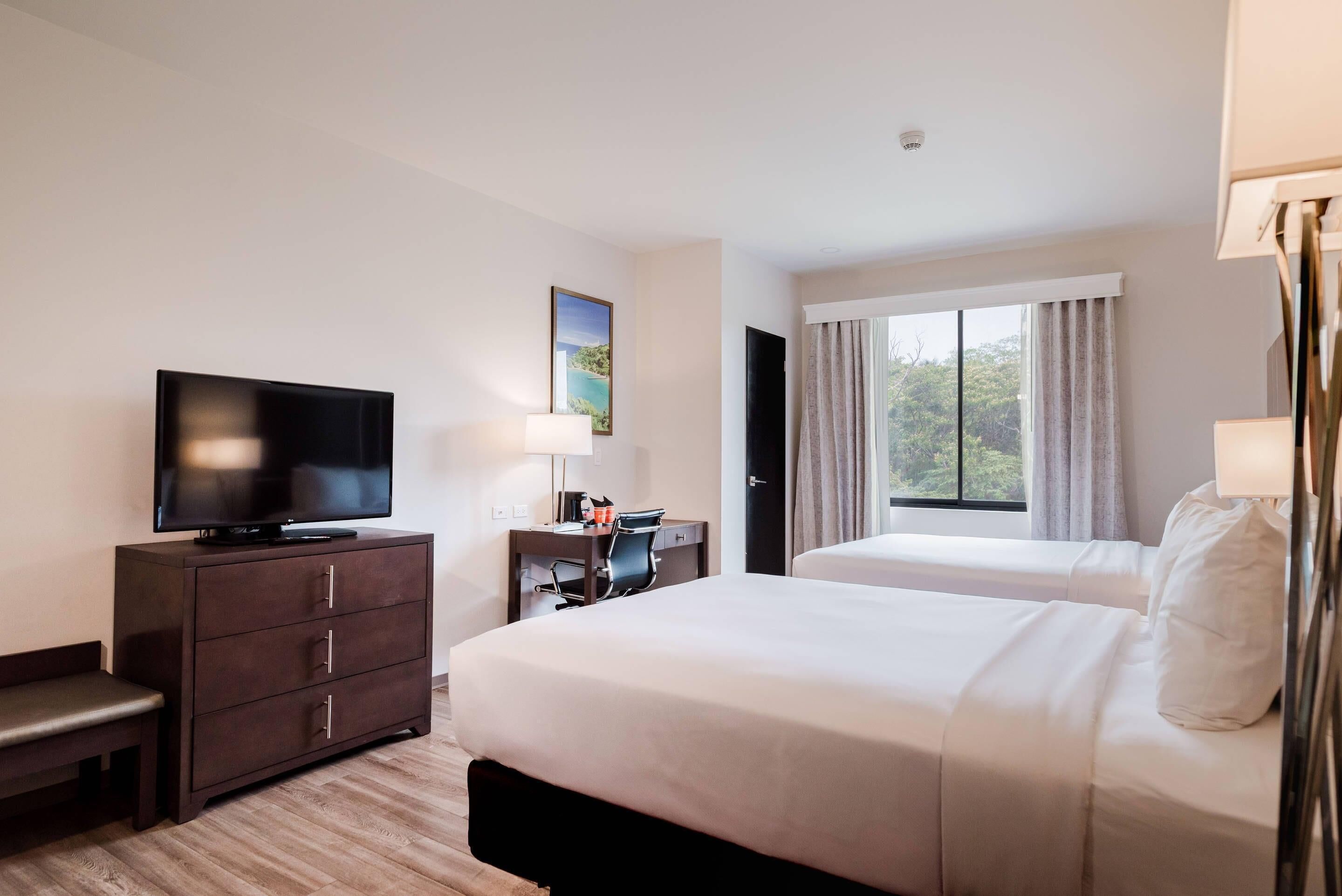 A hotel room with a bed, a desk with a chair, a television, and a window with curtains.