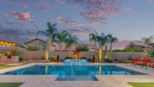 Una piscina rettangolare con bordo piastrellato, circondata da palme e cactus, con un cielo al tramonto sullo sfondo.