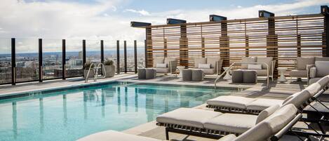 A rooftop pool area with lounge chairs, a wooden slatted wall, and a city view.