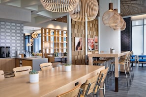 A modern, well-lit dining area with wooden tables and chairs, a central island with a plant, and a seating area with a sofa and bookshelves.