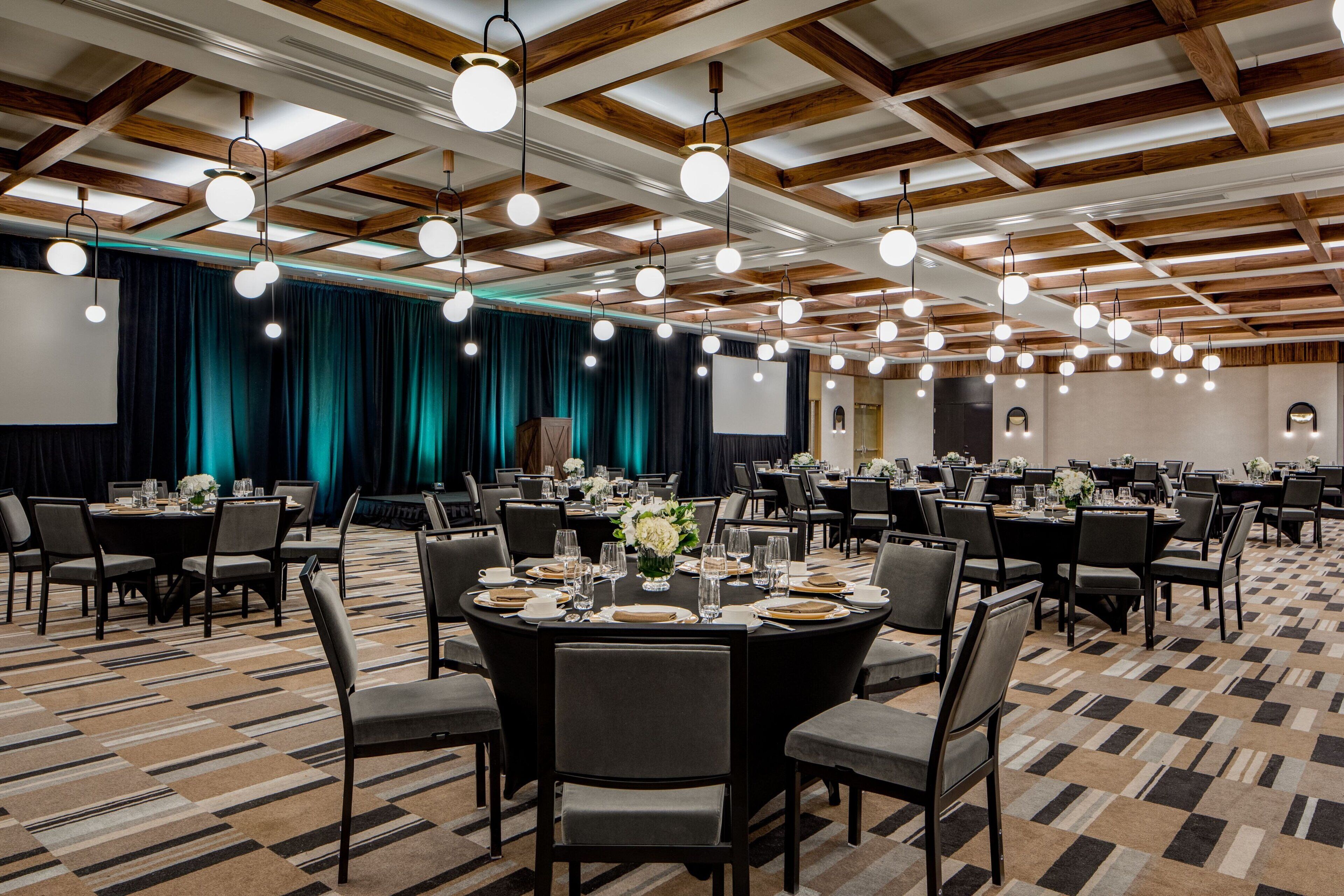 Een ruime feestzaal met ronde tafels gedekt voor een diner, houten balken in het plafond en een podium met gordijnen.