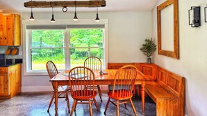 A dining area with a wooden table and chairs, a bench, and a large window with a view of greenery. - Cozy Cabin with Outdoor Shower, Fireplace, Games, Firepit and a Mountain View! (Campton)