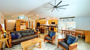 A spacious living area with a black leather sofa, wooden chairs, a wooden coffee table, and a ceiling fan. - Cozy Cabin with Outdoor Shower, Fireplace, Games, Firepit and a Mountain View! (Campton)