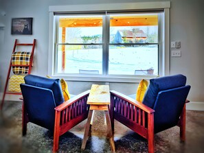 A room with two blue chairs, a wooden table, a window with a view of a building, and a red ladder against the wall.