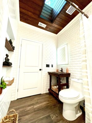 Une salle de bain avec des murs en briques blanches, un plancher en bois, des toilettes blanches, un meuble-lavabo avec un miroir et un ventilateur de plafond.