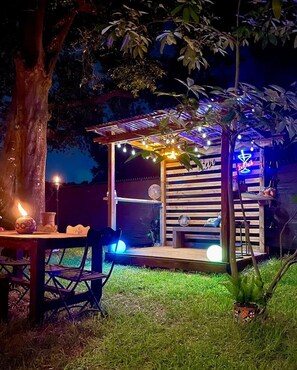 Un espace extérieur aménagé avec une table et des chaises en bois, une petite structure en bois surmontée d’une enseigne au néon et des guirlandes lumineuses.