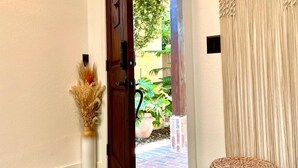 Puerta de madera abierta que da a un jardín, un taburete redondo tejido y una cortina con textura.