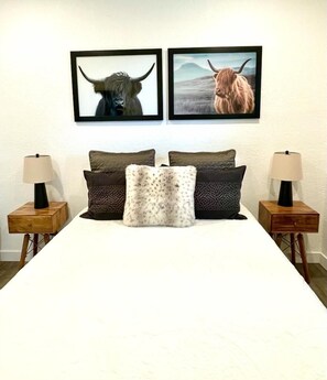 Un lit avec un drap blanc, deux oreillers marron et un oreiller blanc et moelleux. Il y a deux tables de chevet avec des lampes et, au-dessus du lit, sur le mur, on peut voir des photos encadrées de taureaux.
