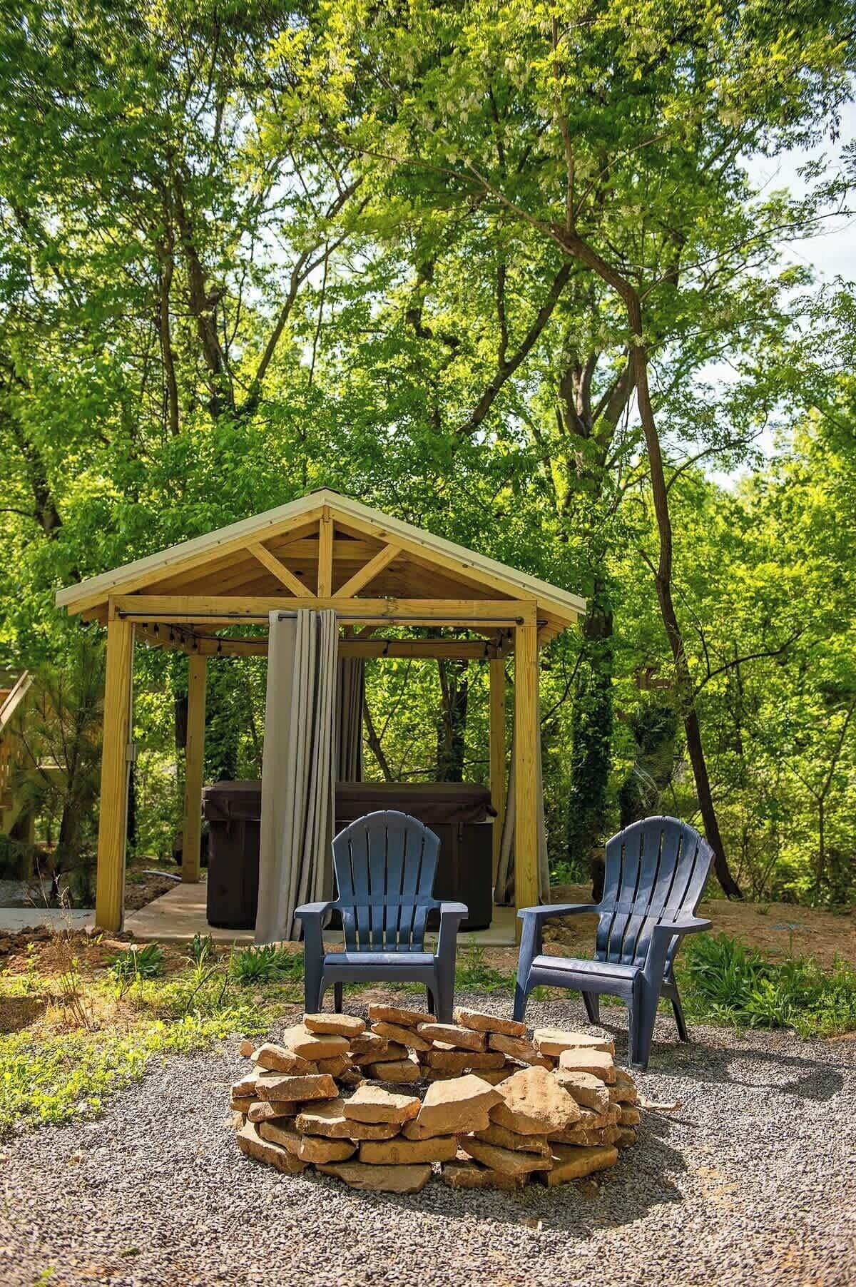 Un belvédère en bois, agrémenté de deux fauteuils Adirondack et d’un foyer en pierre, situé dans un espace boisé.