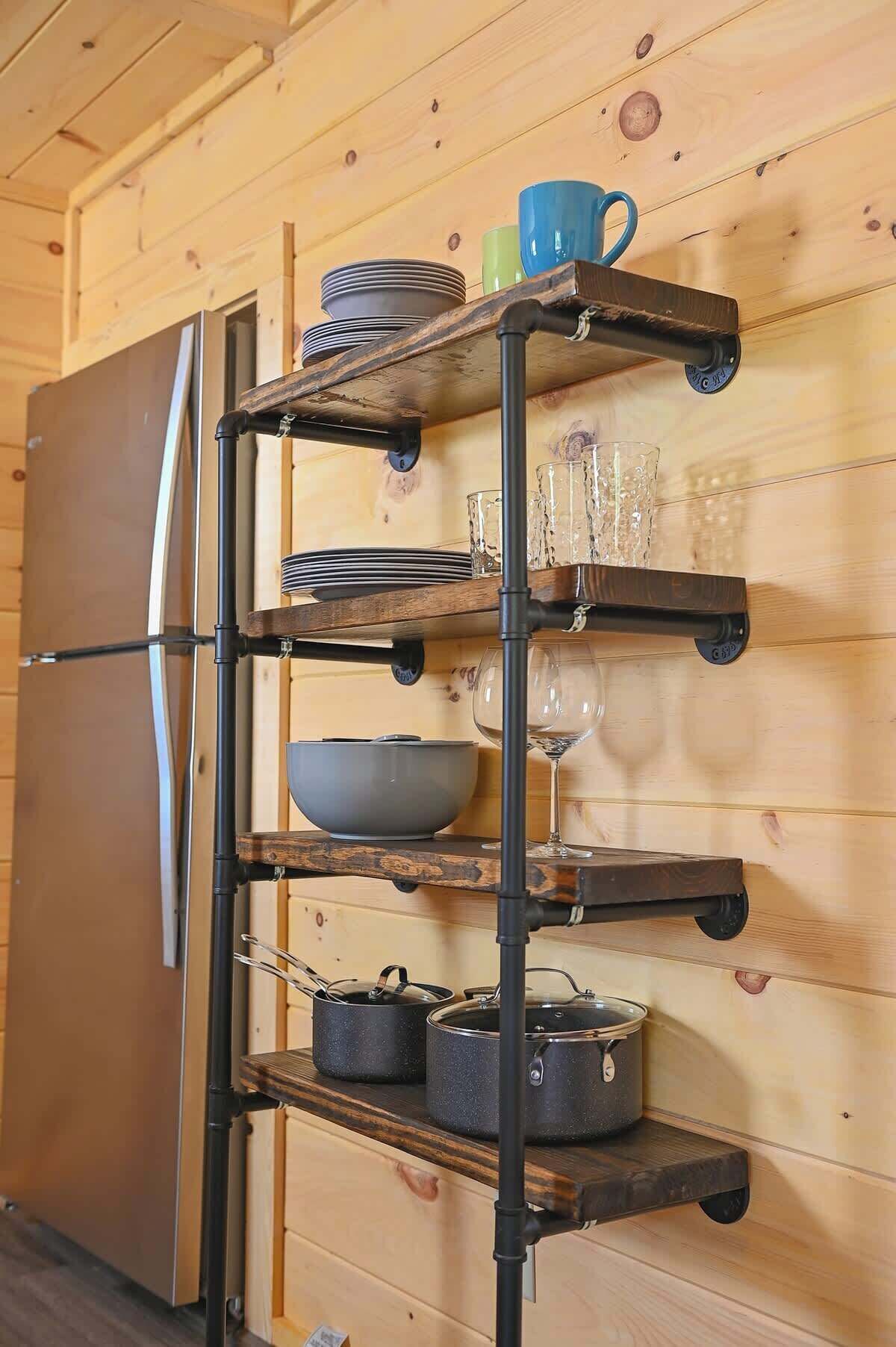 Een keuken met een houten wand, een roestvrijstalen koelkast en een metalen rek met servies, glazen en pannen.