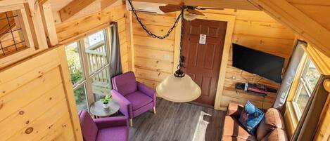Interior acogedor de una cabaña con ventilador de techo, televisor, asientos morados y marrones, y una pequeña mesa con una planta.