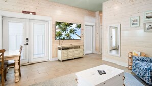 Een ruime woonkamer met een groot flatscreen televisietoestel aan de wand, een houten kastje eronder en een witte salontafel met afstandsbedieningen.