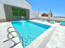 Piscina com água azul cristalina, escada de metal e várias espreguiçadeiras com almofadas azuis.